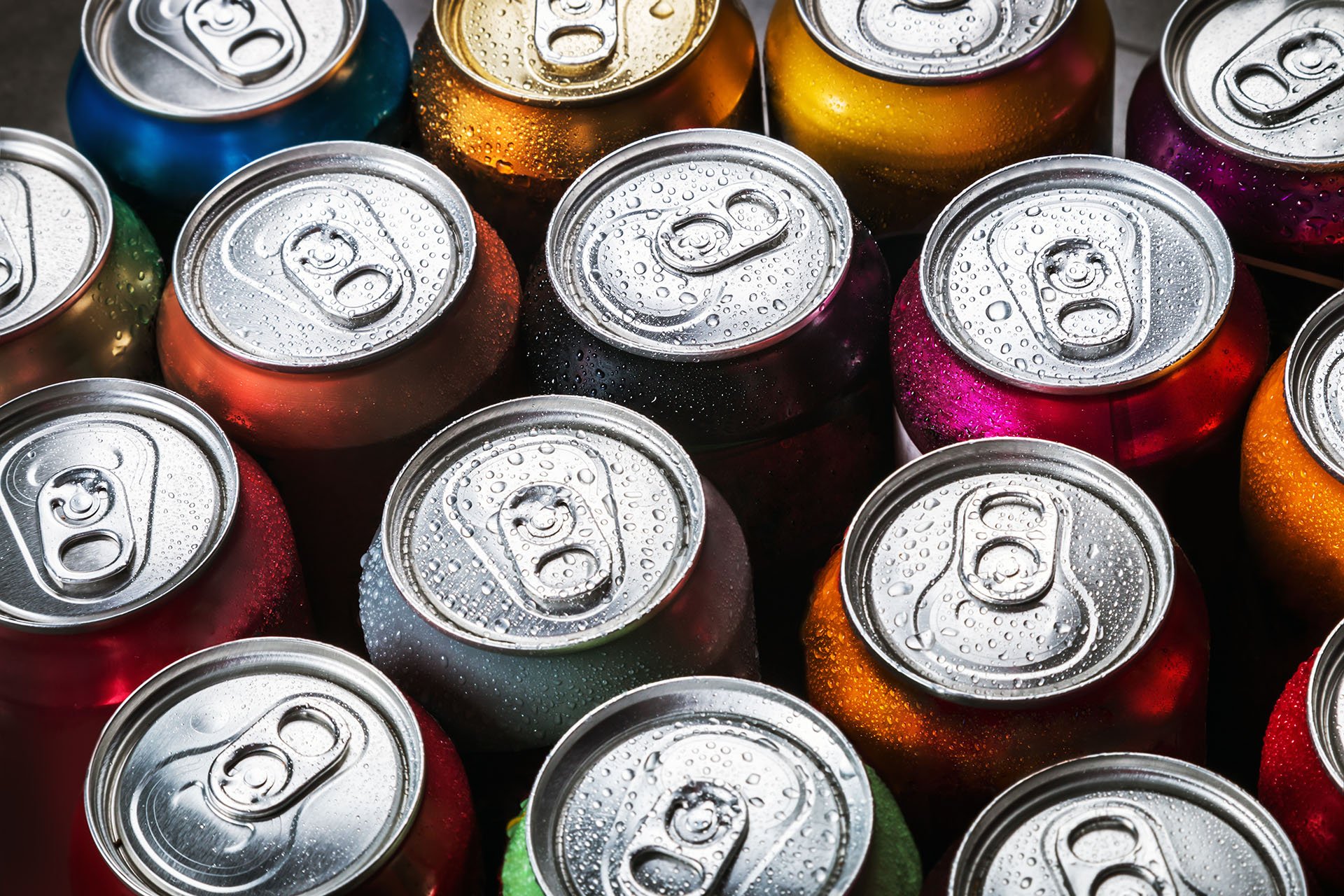 Nærbillede af forskellige sodavandsdåser af aluminium med trækflige, tæt pakket sammen set oppefra og ned. Dåserne er i forskellige levende farver, herunder rød, orange, blå, grøn, lilla og guld, og de er alle dækket af kondensdråber, hvilket indikerer, at de er kolde og friskkølede.