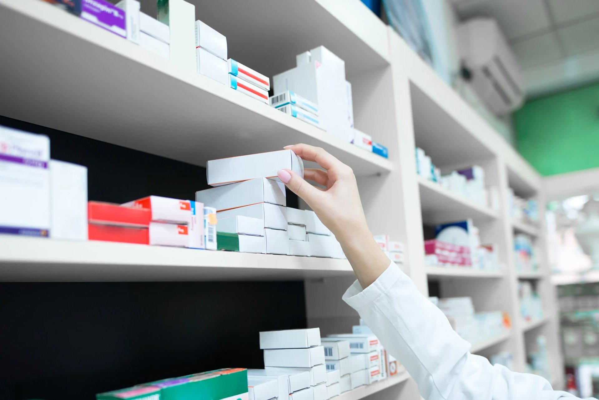 Farmaceutens hånd rækker ud efter en medicinæske på en hylde fyldt med forskellige lægemiddelpakninger.