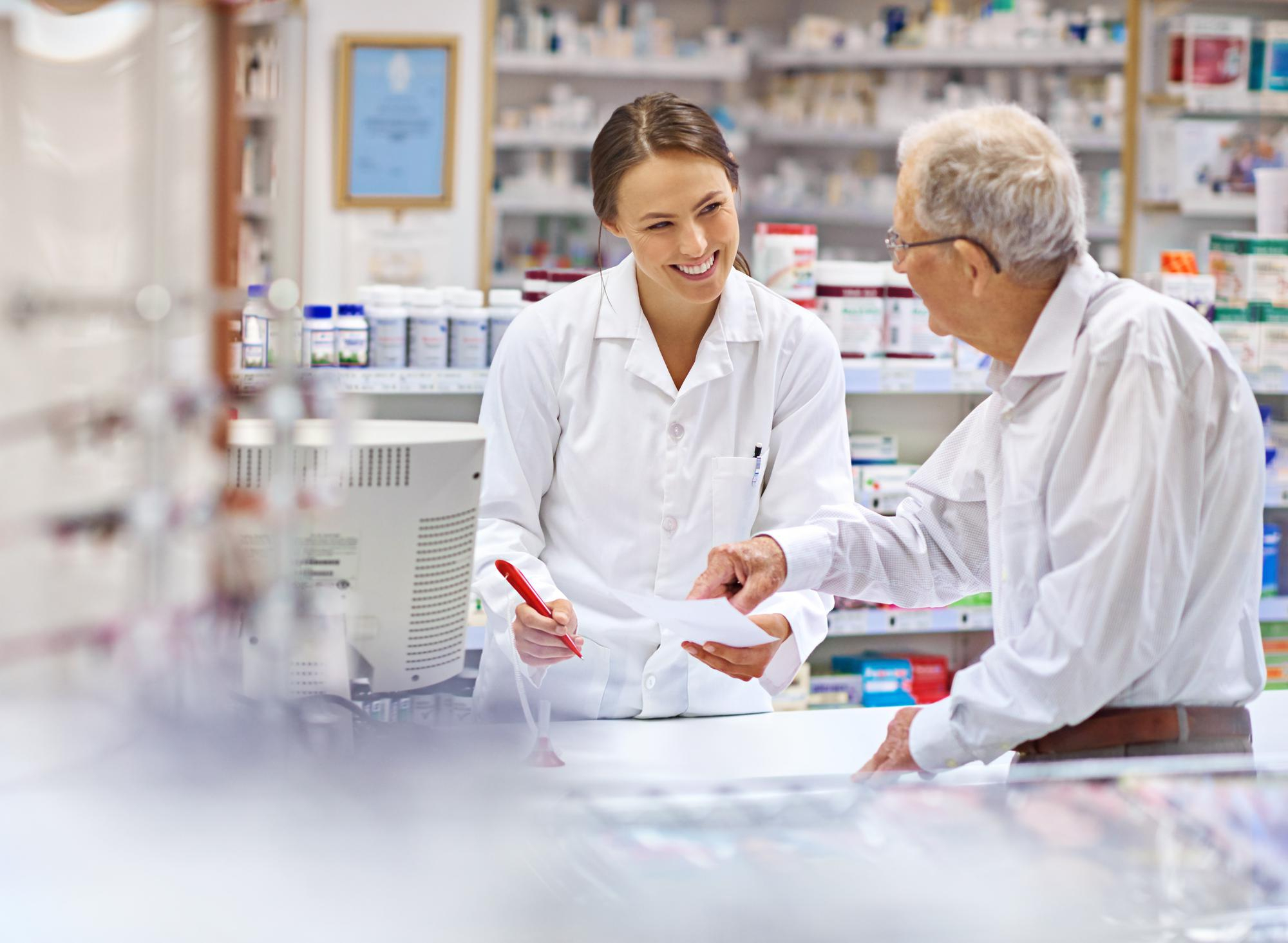 Kvindelig farmaceut hjælper en ældre mandlig kunde med en recept ved en apoteksdisk.