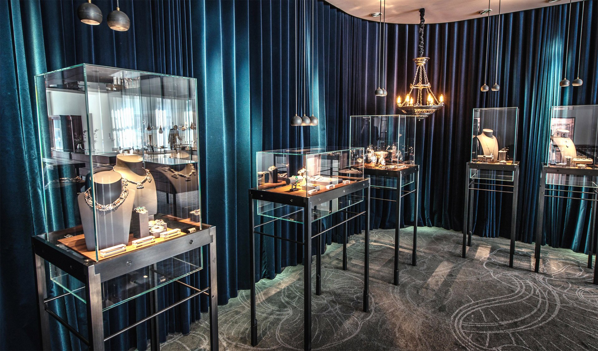 Udstilling af luksussmykker i et svagt oplyst showroom med blå fløjlsgardiner og glasmontrer.