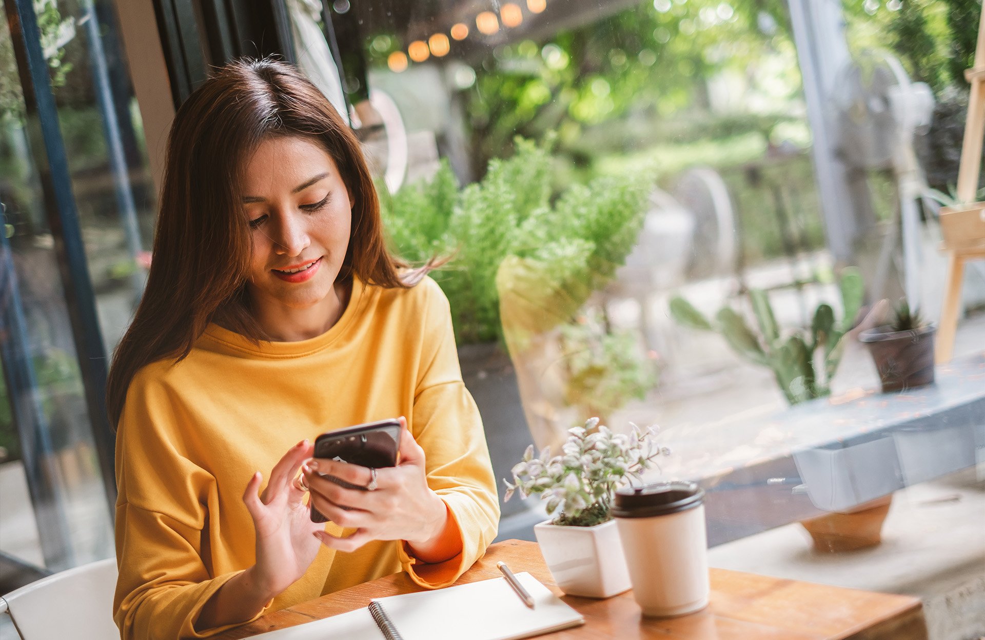 Kvinde bruger en smartphone på en café med en notesbog og en kaffekop på bordet.