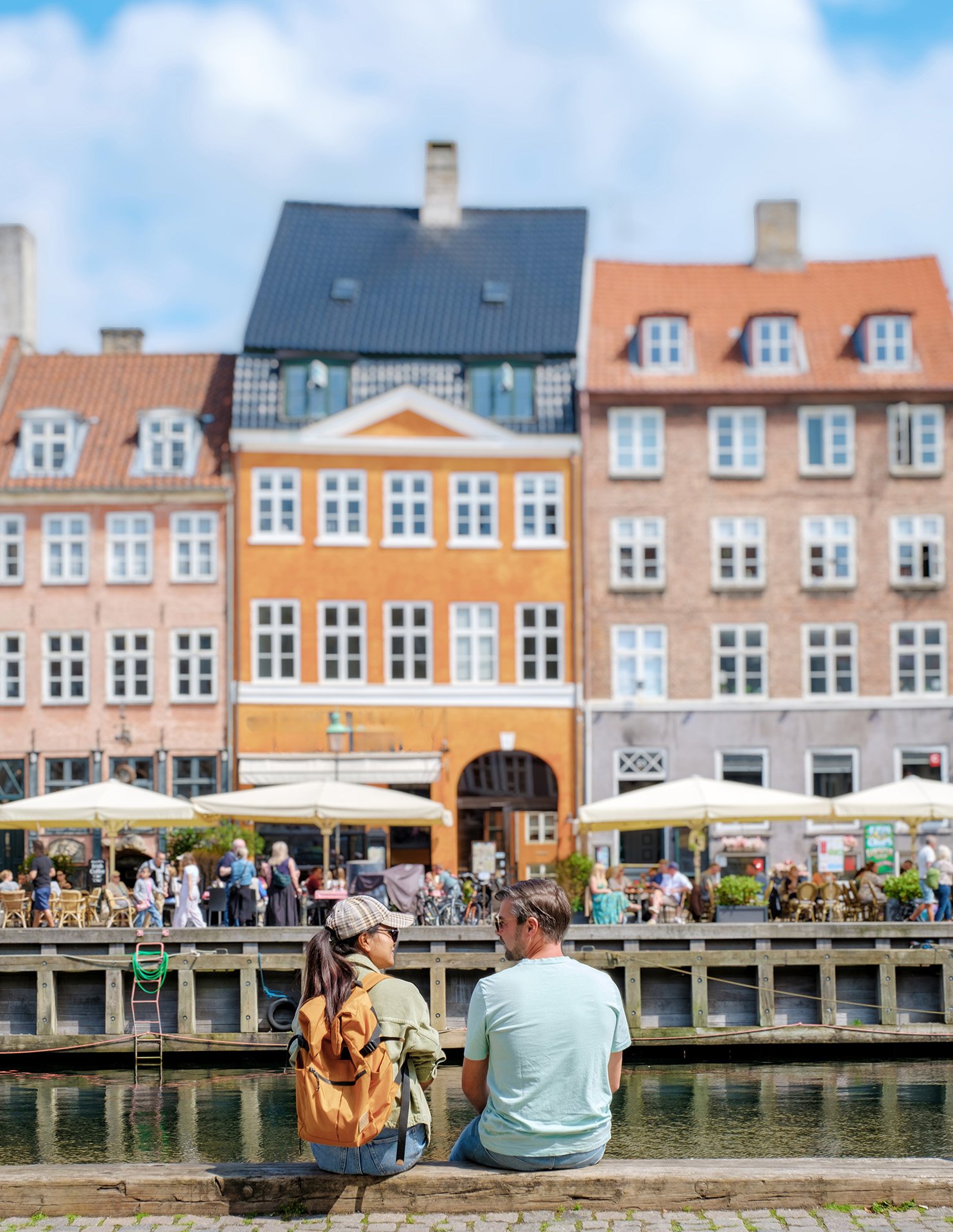 Et par sidder ved en fredelig havnefront i København, omgivet af farverige historiske bygninger og livlige udendørs caféer.
