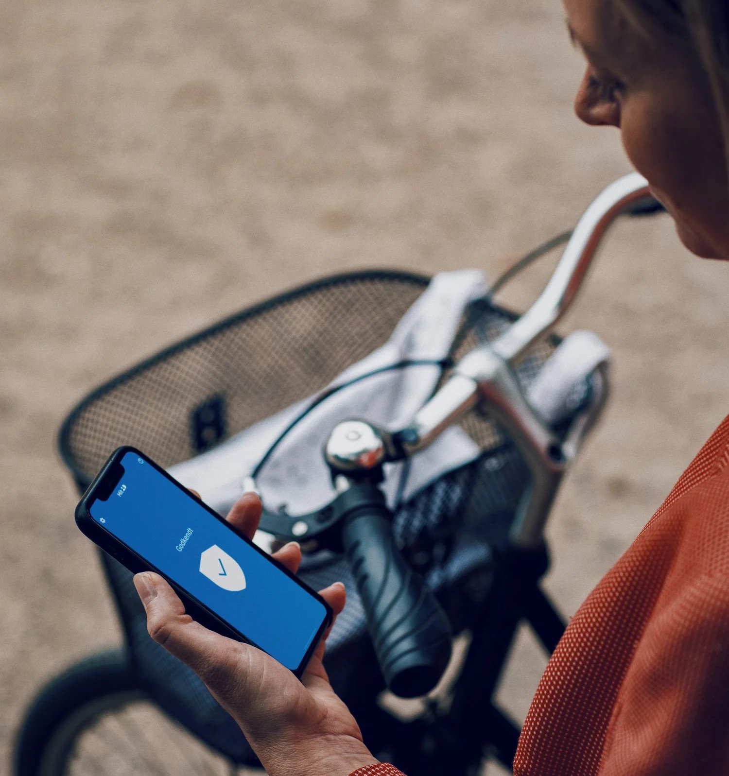 En person holder en smartphone med en blå skærm, der viser et skjold-ikon, ved siden af en cykel med en flettet kurv.