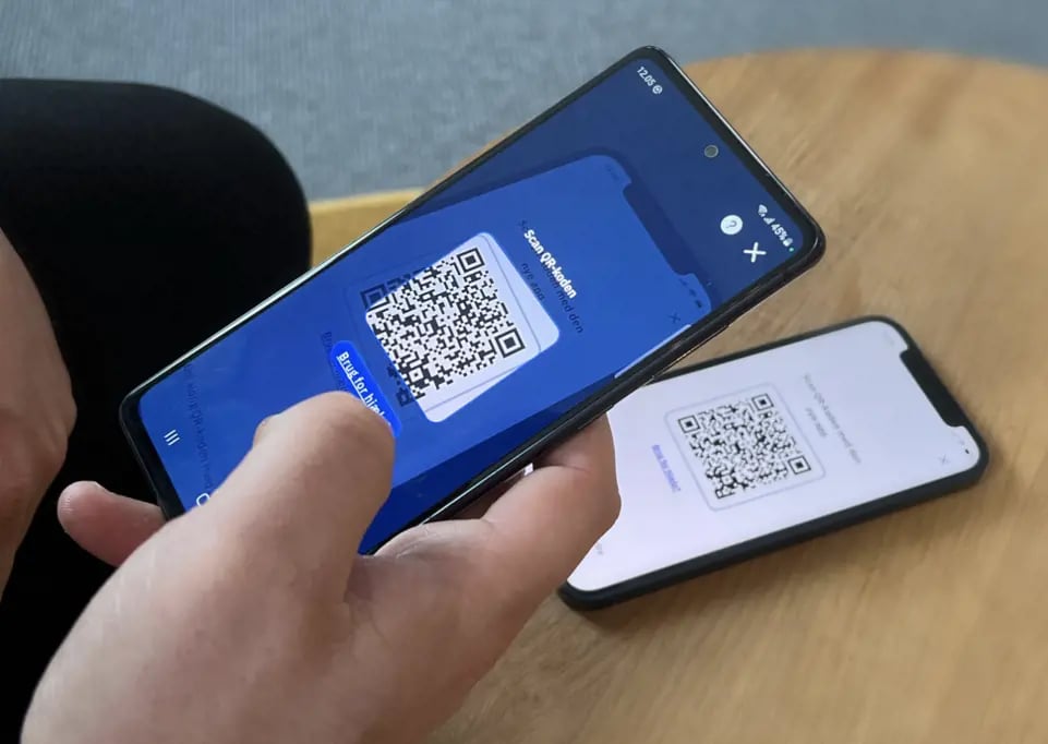 En person holder en smartphone, der viser en QR-kode, mens en anden telefon i baggrunden viser en anden QR-kode på sin skærm.